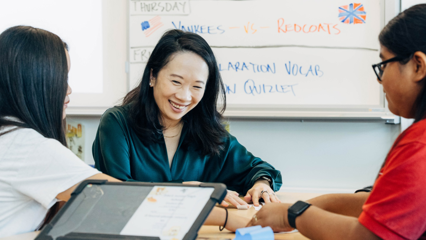 Sandra Liu Huang wears a green blouse and smiles while sitting at a table with three students, engaged in a discussion in a classroom with a whiteboard in the background.