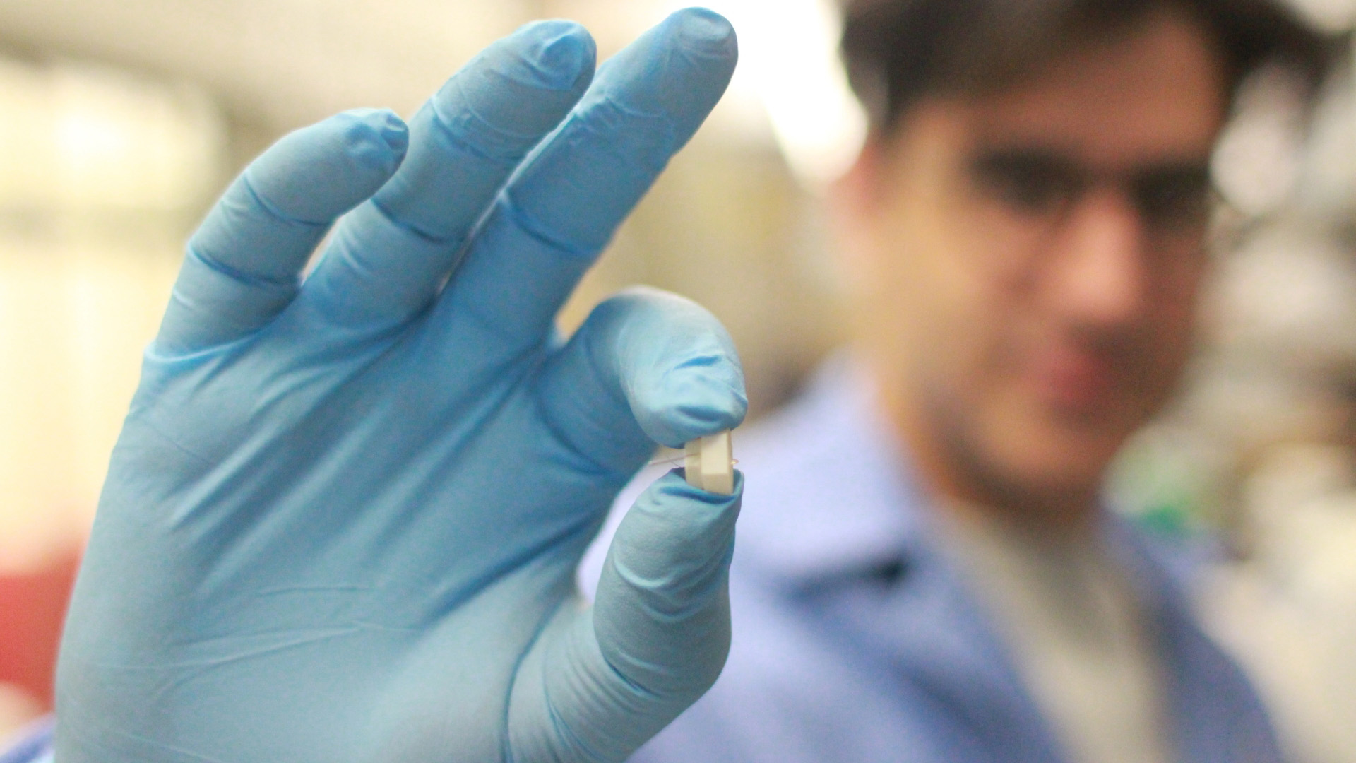 An out-of-focus scientist wearing an in-focus blue surgical glove holds a tiny implantable device between the thumb and index finger.