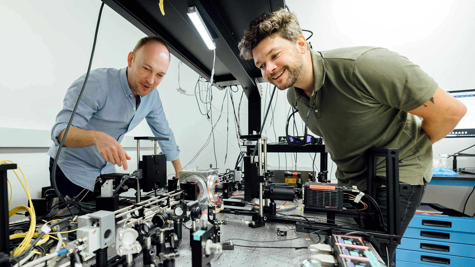 Two researchers examine a complex microscope setup in a laboratory, smiling and engaged, with intricate wiring and equipment on the table in front of them.
