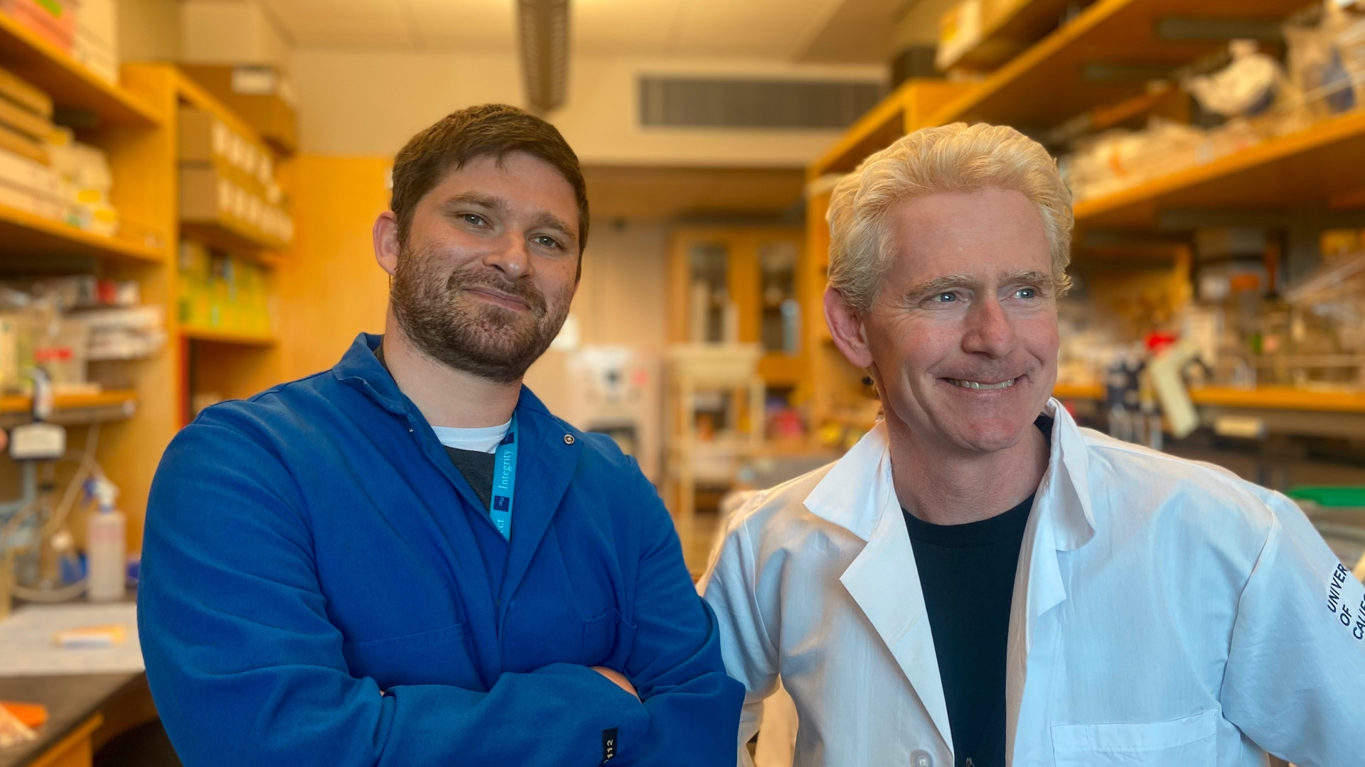 Two researchers stand in a laboratory, smiling, with shelves of scientific equipment and materials in the background. One wears a blue lab coat, and the other a white coat.