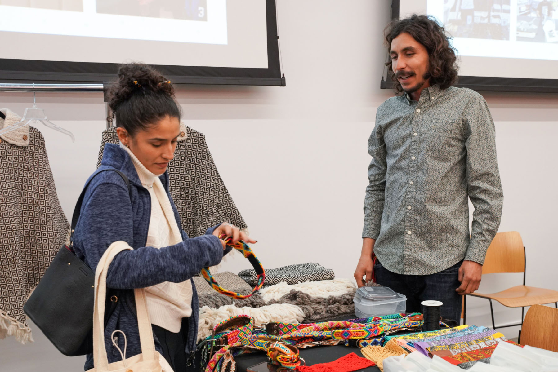A person shops and speaks to a business owner at a table display with handmade jewelry and accessories.