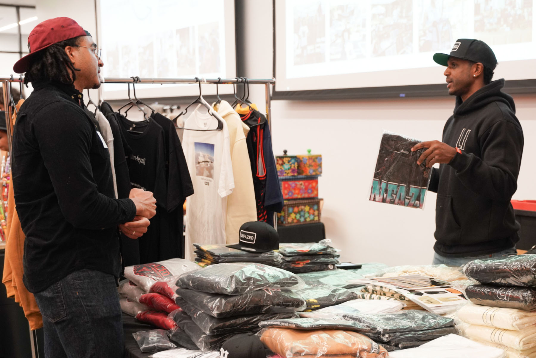 Two men talk over a table display of t-shirts.