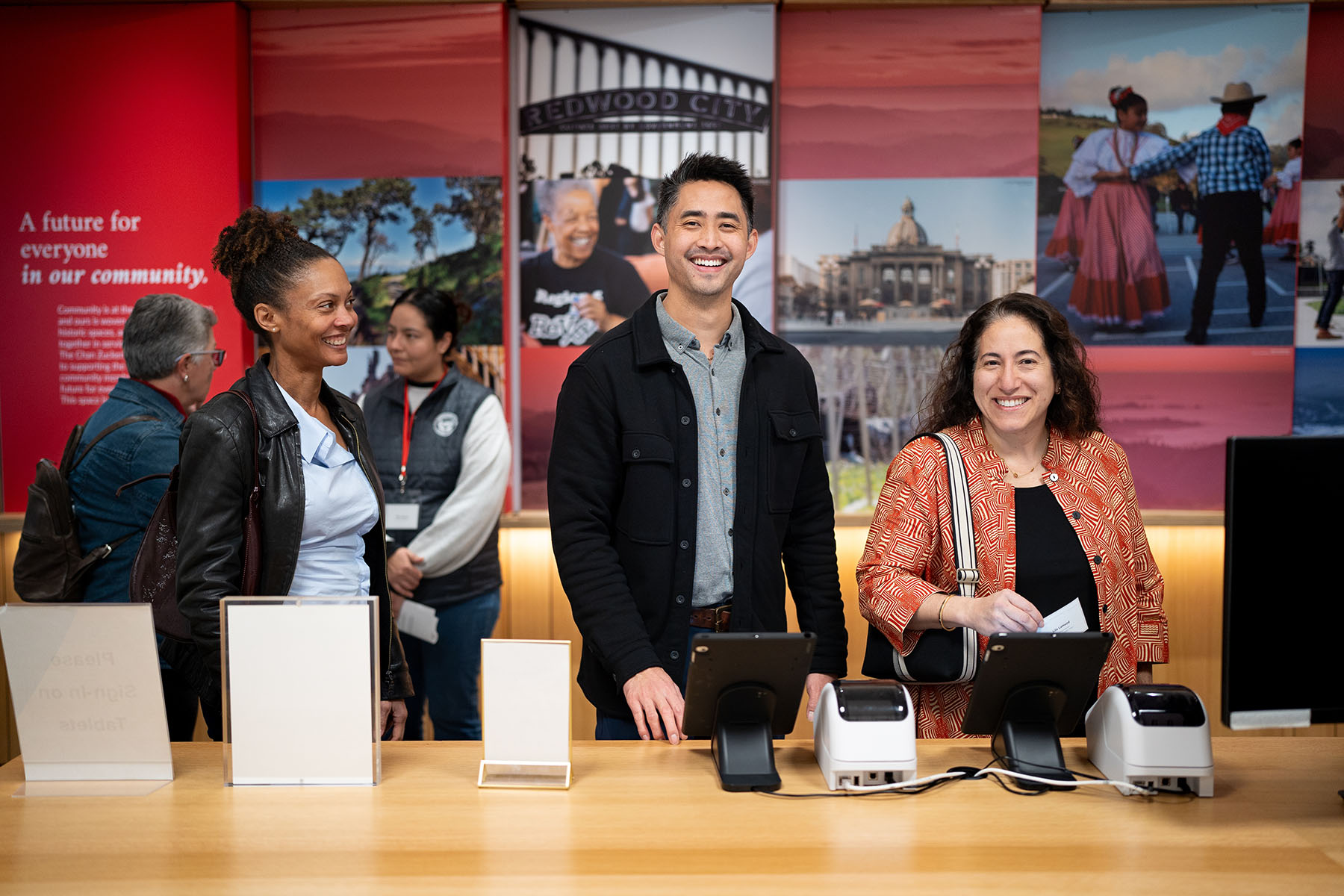 Two people smile at the camera as they register while a person to the left smiles at them.
