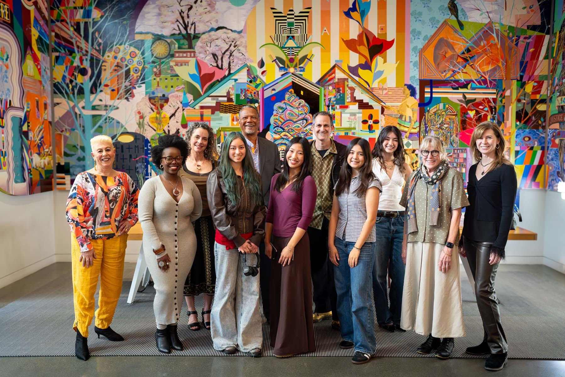 Eleven people stand closely together and smile in front of a colorful mural.