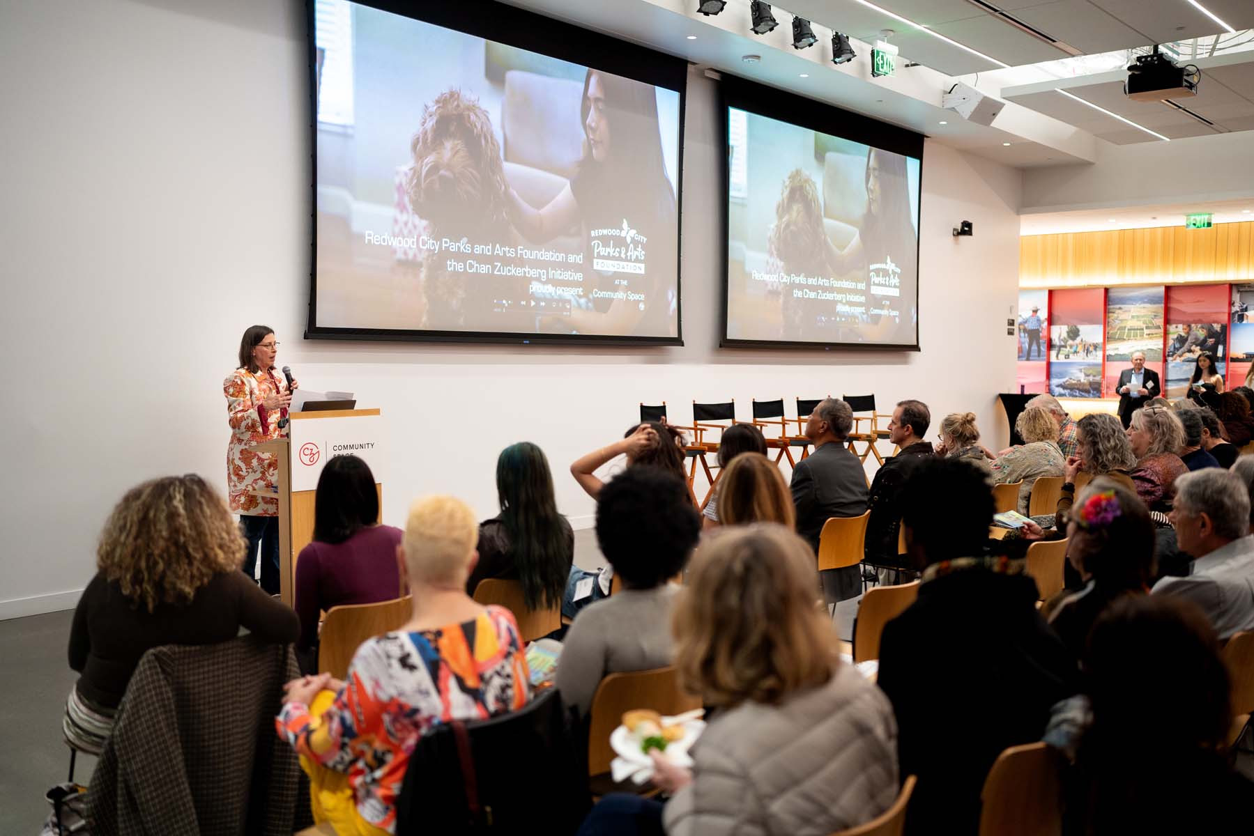 A woman holding a microphone stands at a podium before a small crowd of people who are seated.