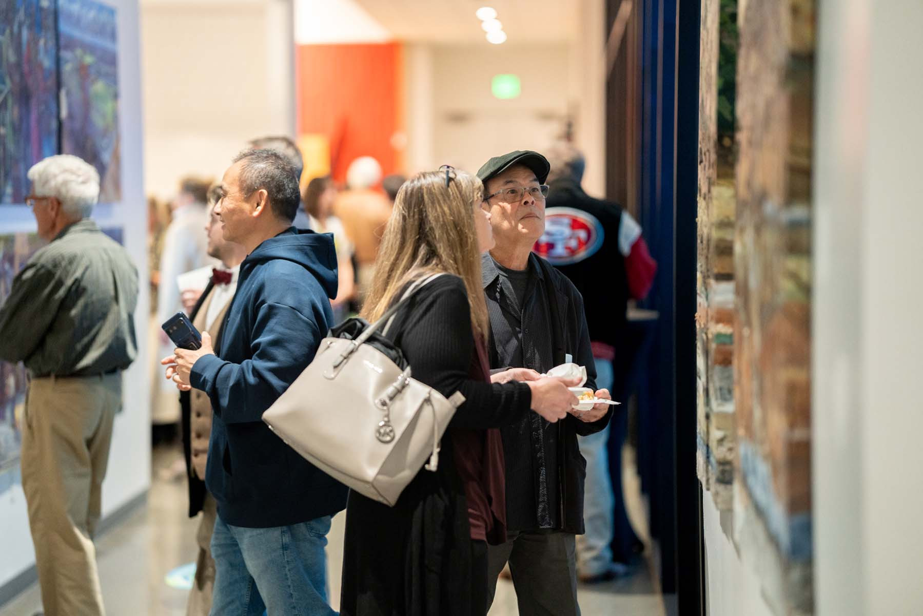 A couple stands together to look at art on a wall as others do the same in the background.