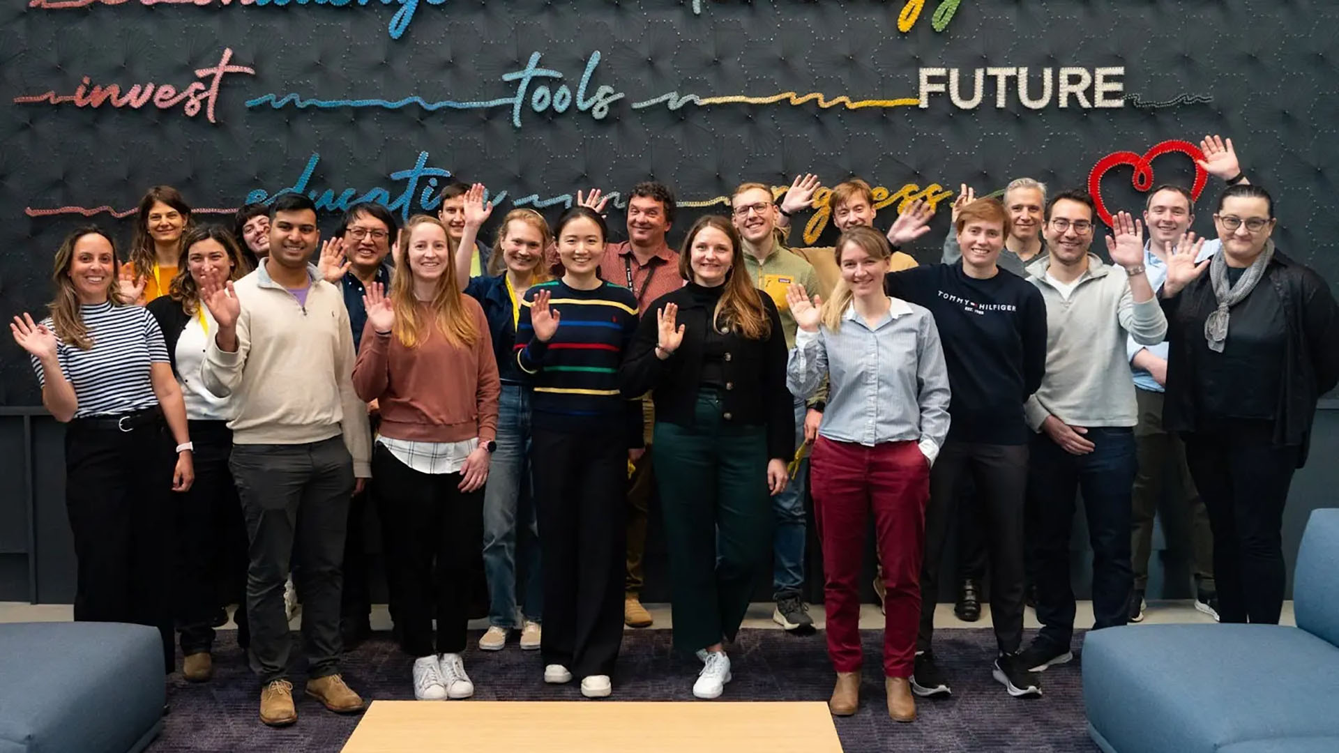 Single-cell community benchmarking working group photo of smiling people waving at the camera in front of a wall with colorful text including the words “technology,” “tools,” and “future.”